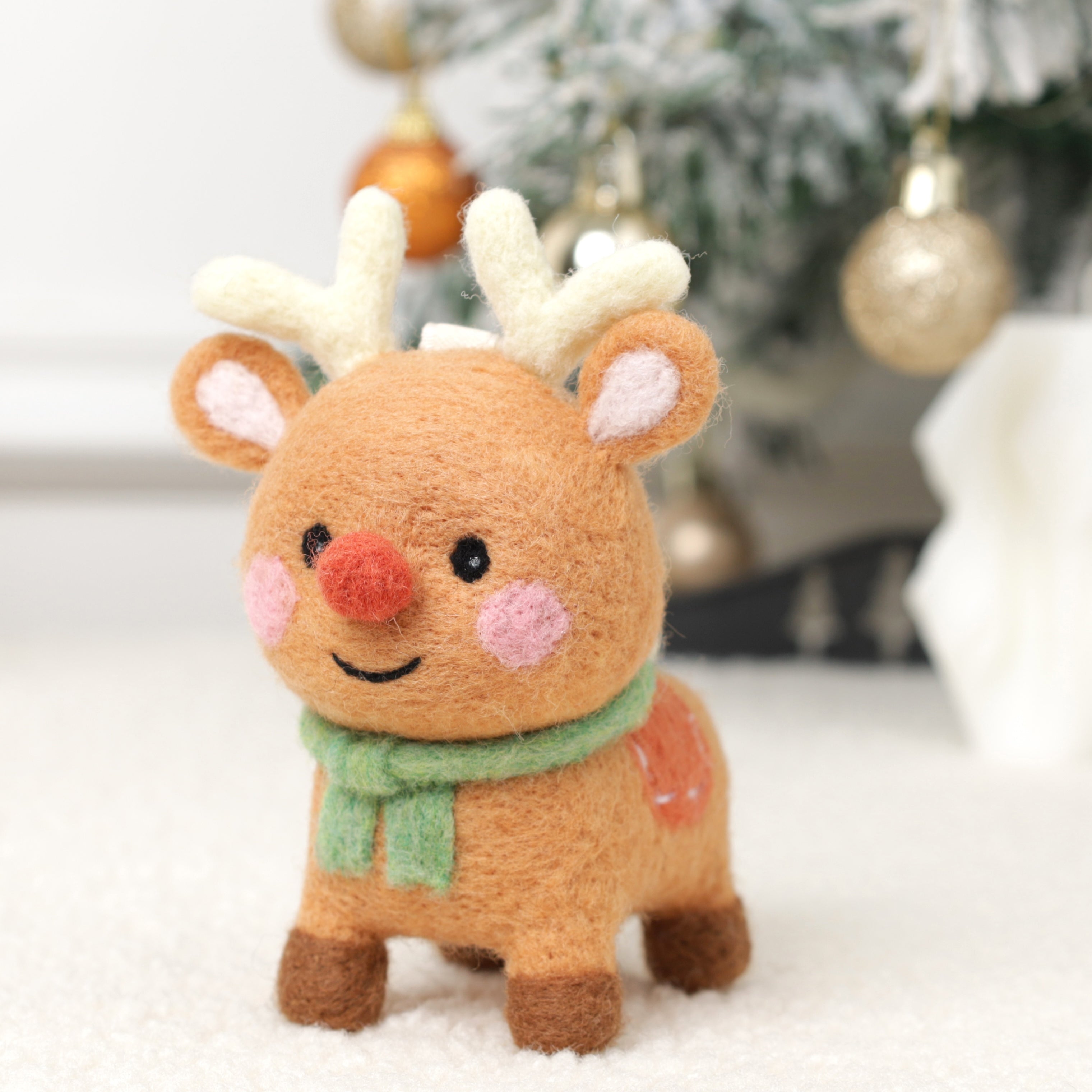 Handmade Merino wool Woobbi small reindeer with a green scarf in front of a Christmas tree.