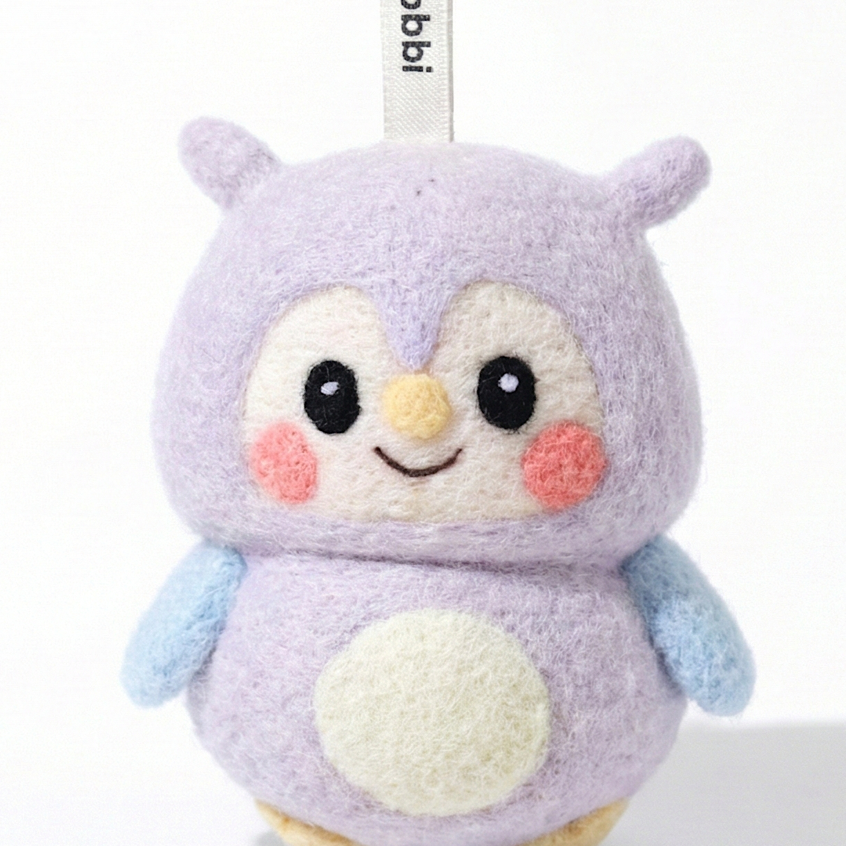 Front view of a handmade Woobbi owl keychain, crafted from soft lavender wool felt. The owl features large black eyes, rosy pink cheeks, a small yellow beak, and a cream-colored belly patch. It has light blue wings and is attached to a black metal keyring with a white tag labeled 'Woobbi'. The background is plain white with soft studio lighting.
