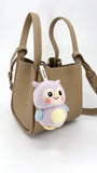 Handcrafted needle-felted wool bag charm featuring a cute purple owl design, styled on a beige leather bucket bag to show size and texture.